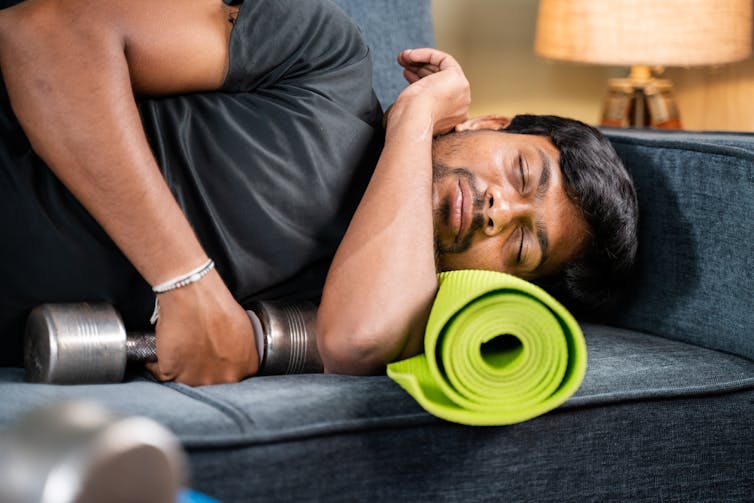A tired man sleeps on his couch using a yoga mat for a pillow and holding a dumbbell.