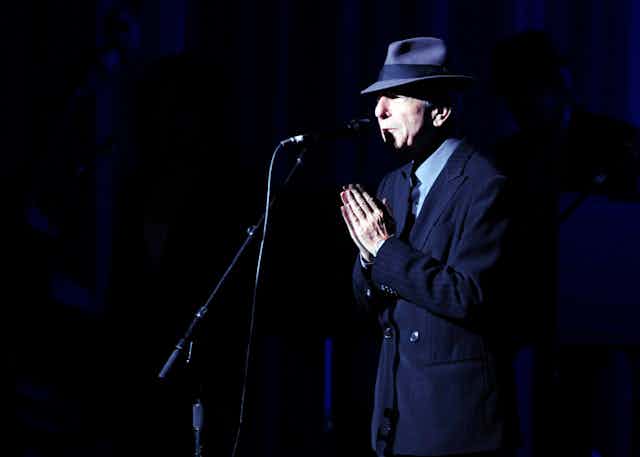 A man at a microphone in a fedora with hands folded in gesture of gratitude.