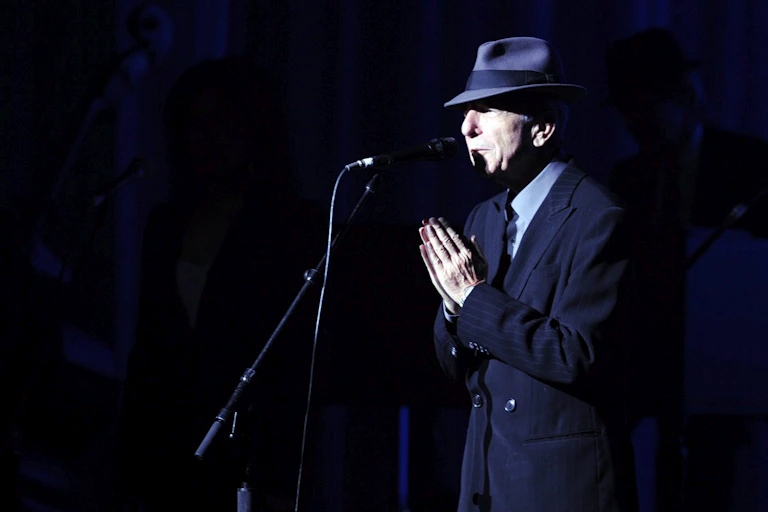 A man at a microphone in a fedora with hands folded in gesture of gratitude.