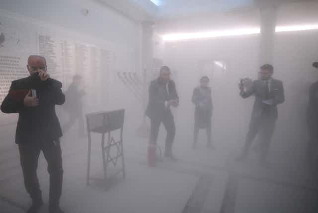 Grzegorz Braun standing in a foggy room over a fire extinguisher with a Hanukkah menorah behind him, its candles blown out. Other people are moving around him 