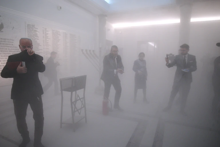 Grzegorz Braun standing in a foggy room over a fire extinguisher with a Hanukkah menorah behind him, its candles blown out. Other people are moving around him