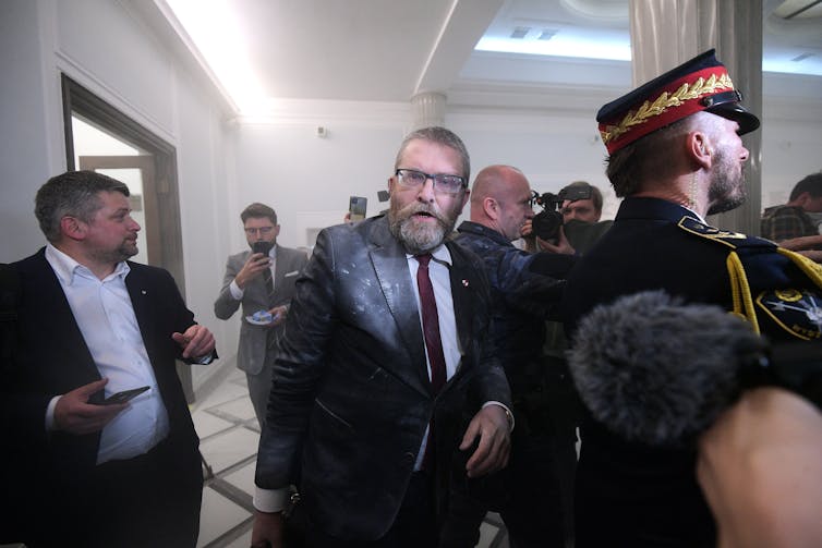 Grzegorz Braun covered in fire extinguisher residue in the middle of a crowd standing behind a guard.