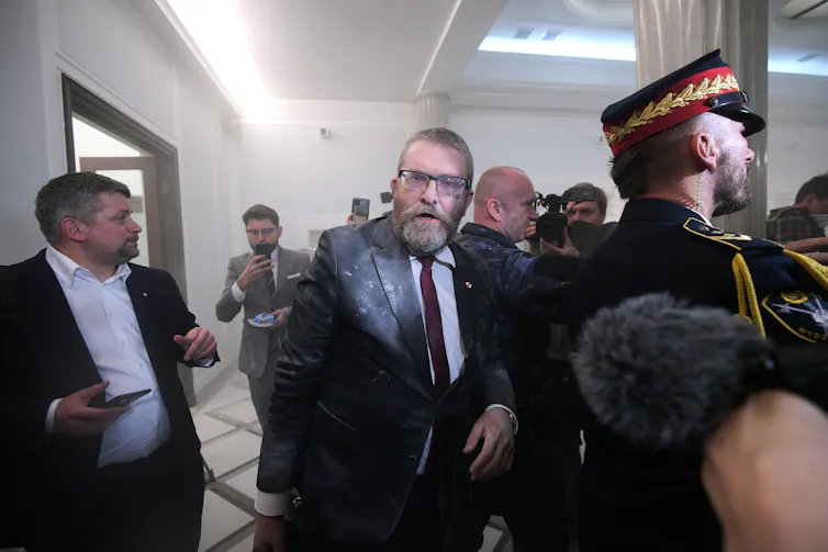 Grzegorz Braun covered in fire extinguisher residue in the middle of a crowd standing behind a guard.