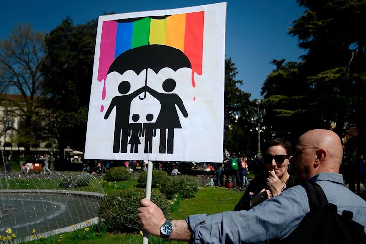 A man holds a placard with stick figures of a man and a woman holding an umbrella over two children below a rainbow-painted sky.