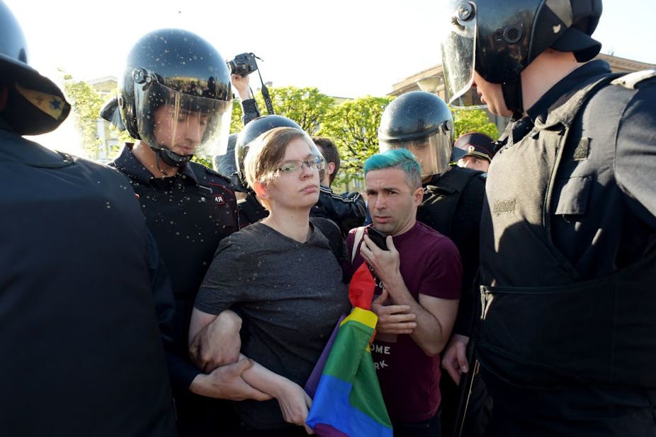 A serious-looking person holding a rainbow flag and a man with blue hair are surrounded by police in black outfits. 