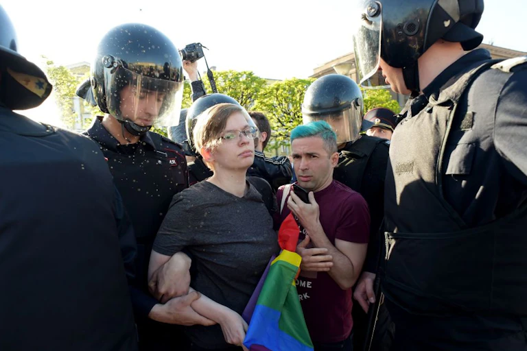 A serious-looking person holding a rainbow flag and a man with blue hair are surrounded by police in black outfits.