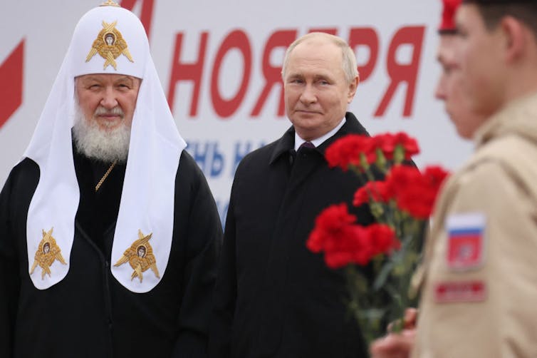 A man with a white beard, in black and white priest's clothing, stands next to a balding man in a black coat and tie.