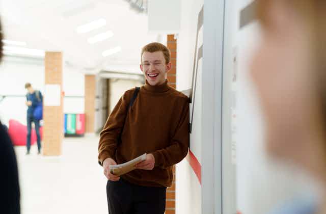 A young man in a backpack learns against a wall, holding papers and smiling. 