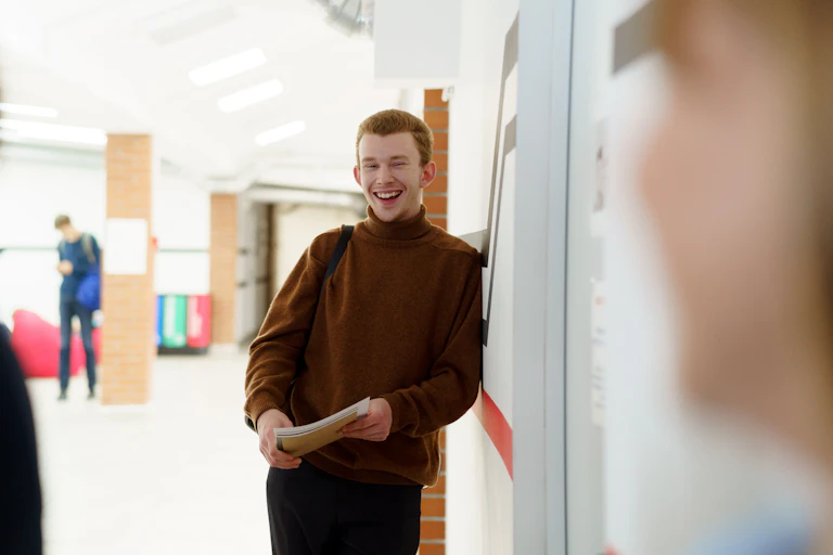 A young man in a backpack learns against a wall, holding papers and smiling.