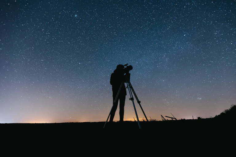 A person silhouetted against a night sky looking through a telescope