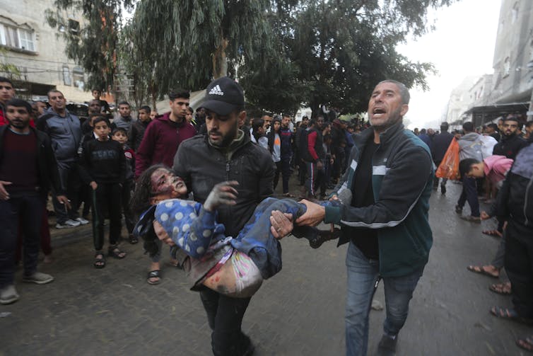 A man carries a wounded young girl while another man beside him sobs.