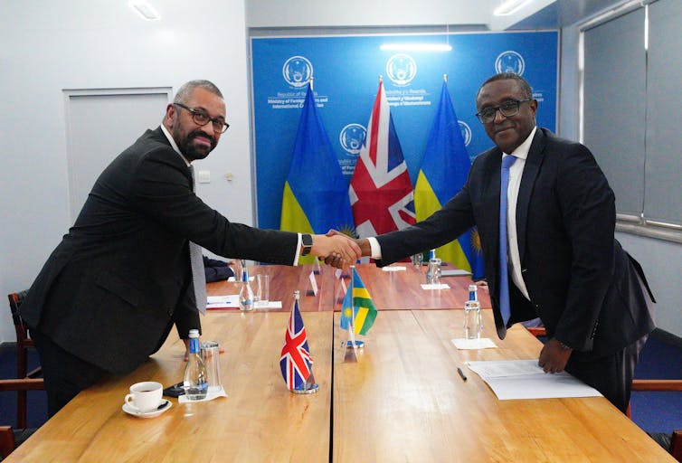 James Cleverly and Vincent Biruta smiling and shaking hands over a conference table featuring small UK and Rwandan flags.