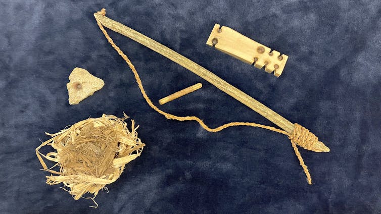 A set of wood tools, including a long stick with a cord attached, a small stick, a piece of wood with grooves carved into it, a pile of dried grass, and a small, triangle-shaped stone.