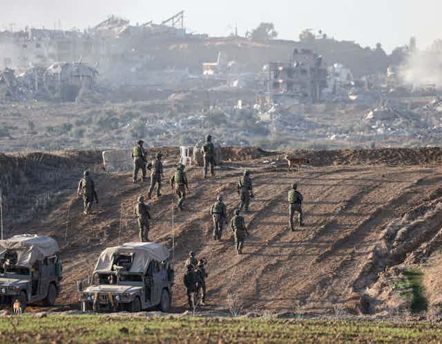 A group of soldiers are walking on a dirt road near buildings that have been destroyed. 