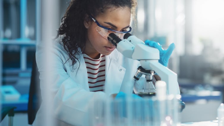 A black woman in a lab coat looking through a microscope
