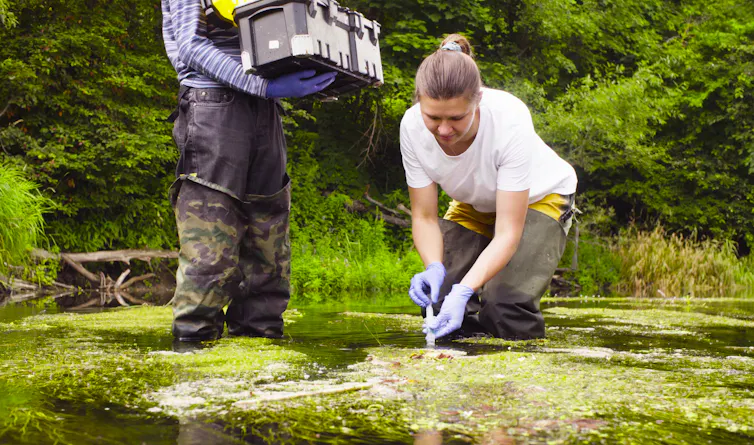 A white woman takes a sample from a body of water. Another person stands next to her carrying a box.