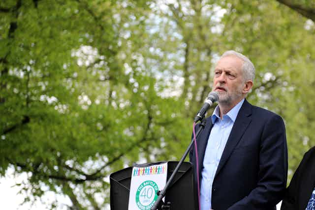 An older man (jeremy Corbyn) in a blue suit making a speech in a park.