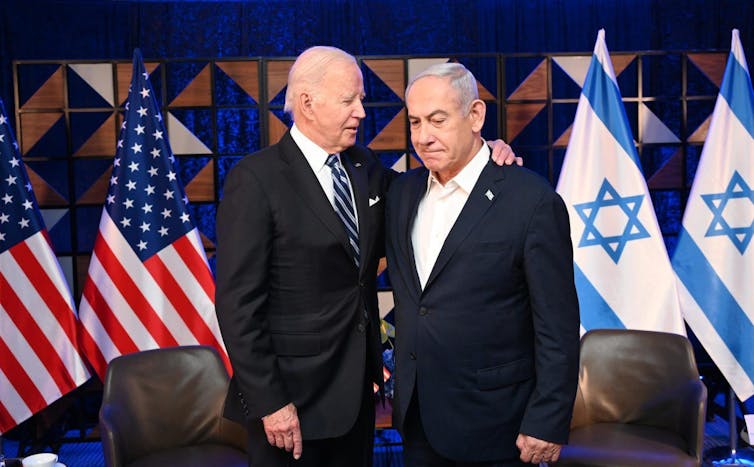 An elderly white man has his arm around another man as both them stand before Israeli and US flags.