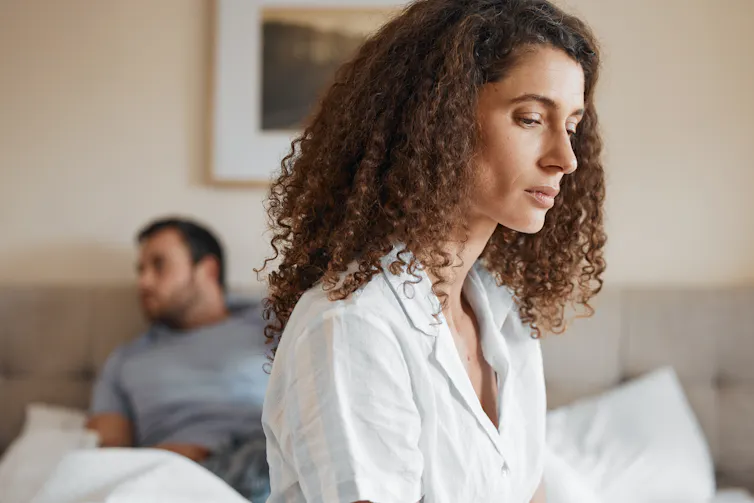 A woman with a sad expression sits on the edge of a bed. A man sits on the other end of the bed behind her.