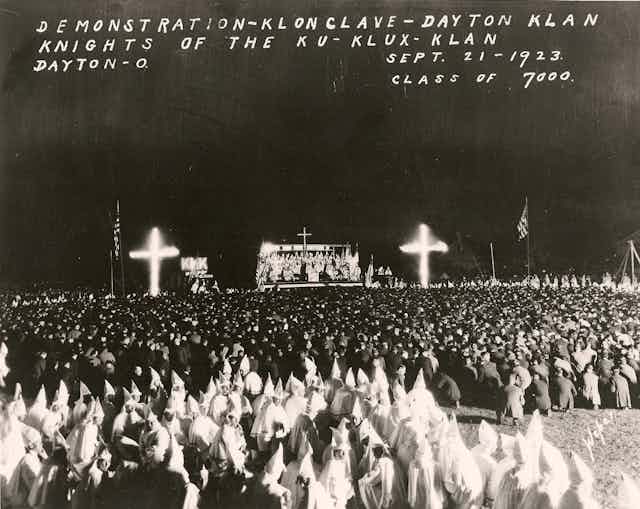 A black and white photograph shows large burning crosses at the front of a large crowd of people, many of them in white robes with hoods.