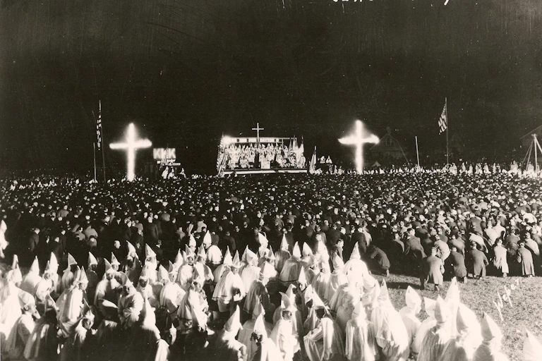 A black and white photograph shows large burning crosses at the front of a large crowd of people, many of them in white robes with hoods.