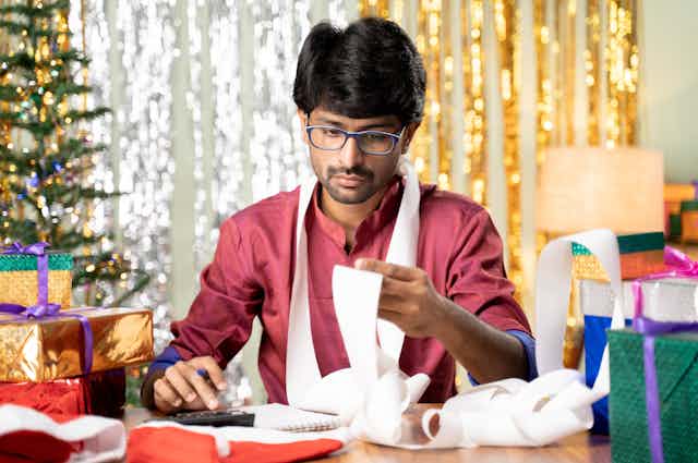 A man sitting at a table surrounded by gift-wrapped presents looking at a roll of receipt paper so long it curls around his neck