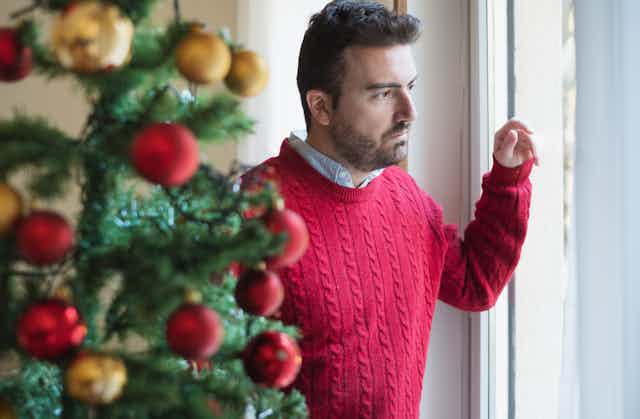 A sad man in a red sweater stands beside a christmas tree looking out a window