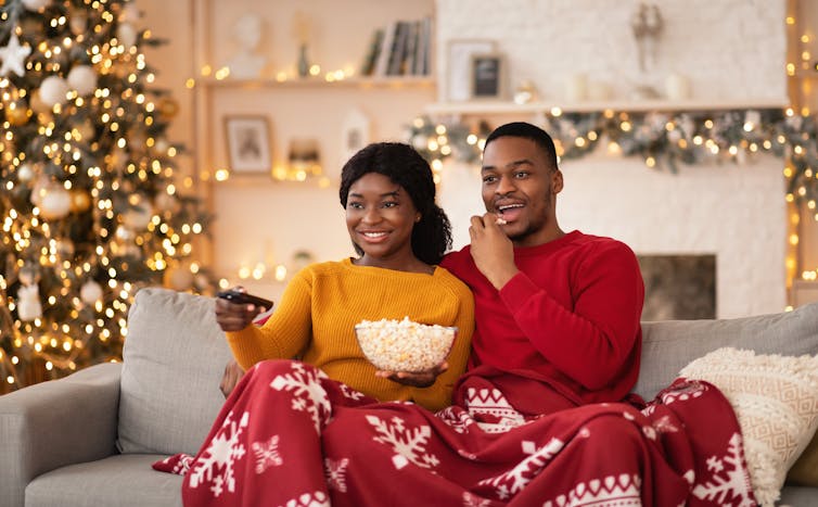 A young Black couple sit on a couch with popcorn watching a movie. A christmas tree and decorations can be seen behind them.
