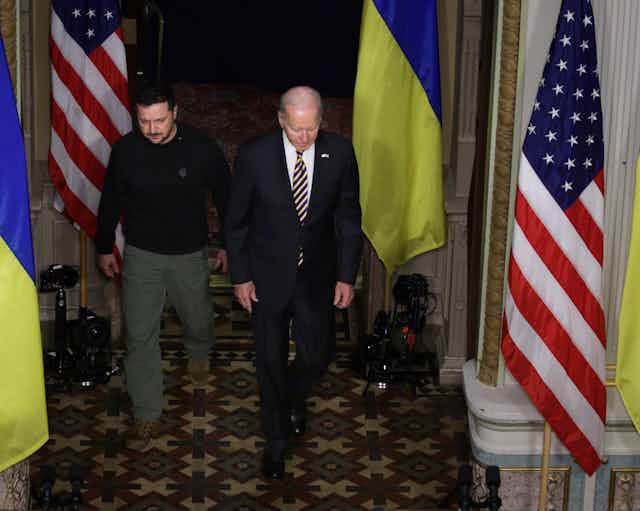 A man dressed in a business suit is walking with another man wearing a dark shirt in a room with two different flags. 