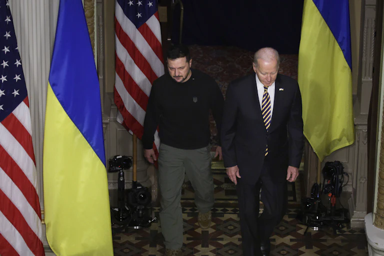 A man dressed in a business suit is walking with another man wearing a dark shirt in a room with two different flags.