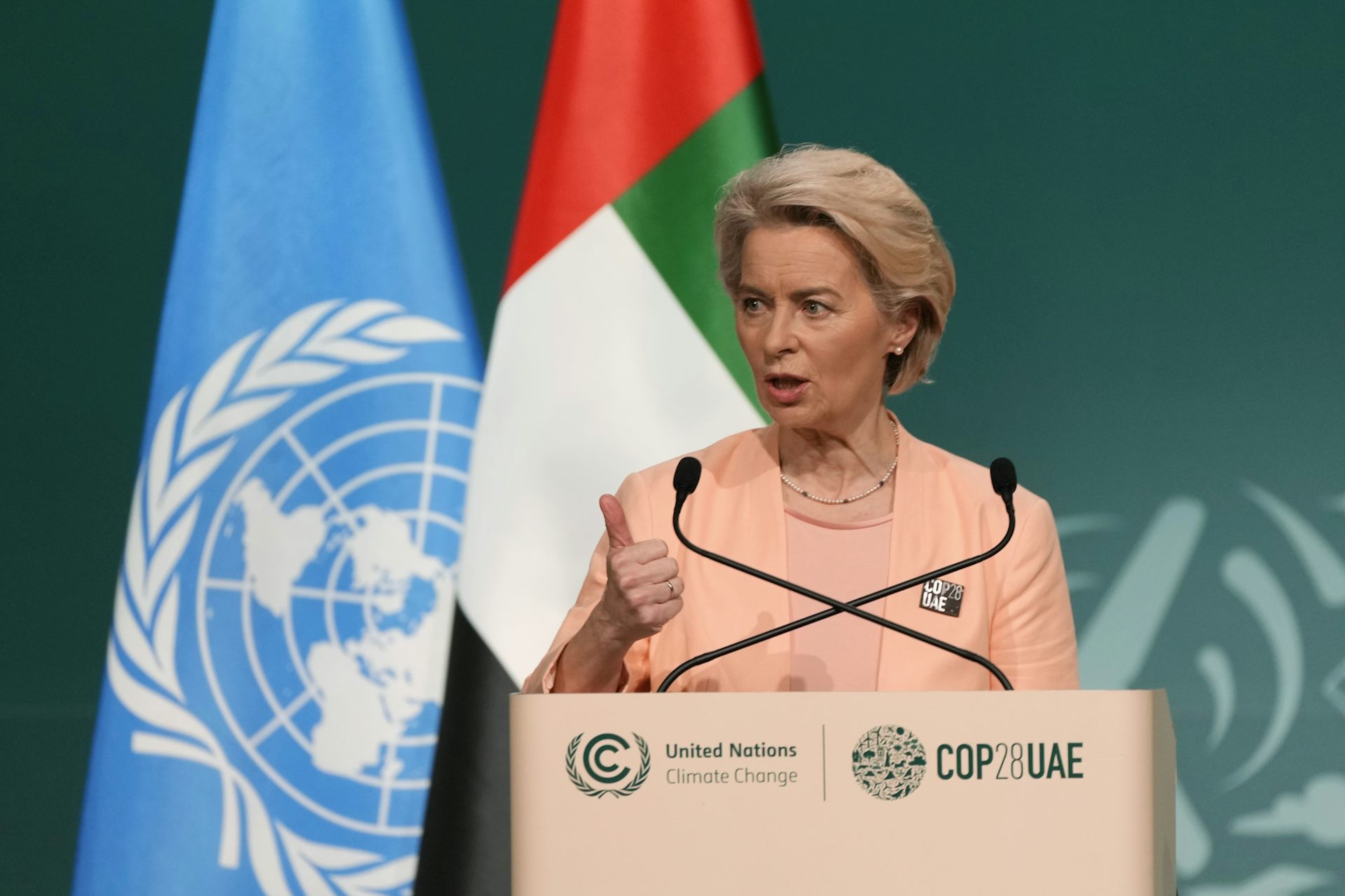 A middle-aged woman with short hair speaks from behind a podium. The UN and United Arab Emirates flags stand behind her.