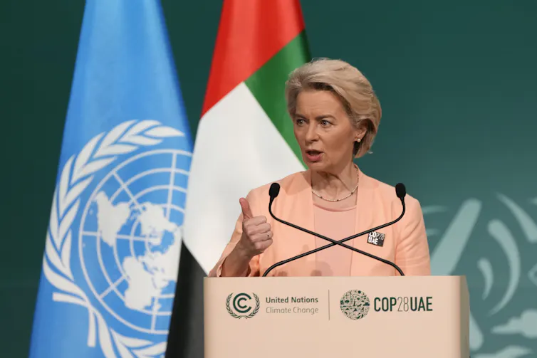 A middle-aged woman with short hair speaks from behind a podium. The UN and United Arab Emirates flags stand behind her.