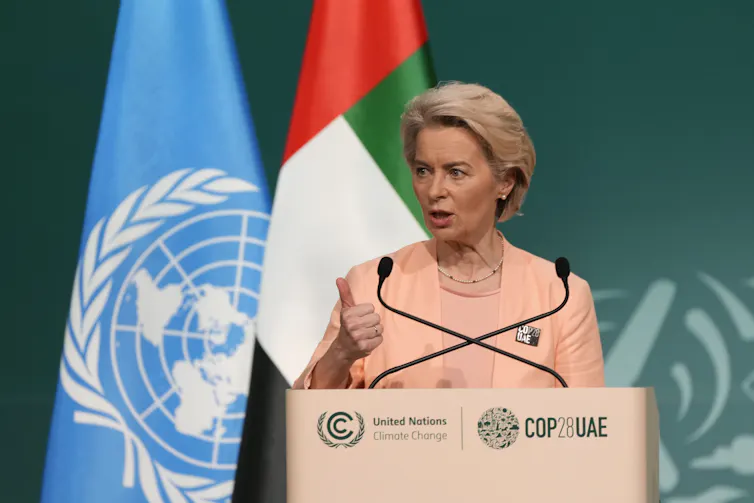 A middle-aged woman with short hair speaks from behind a podium. The UN and United Arab Emirates flags stand behind her.