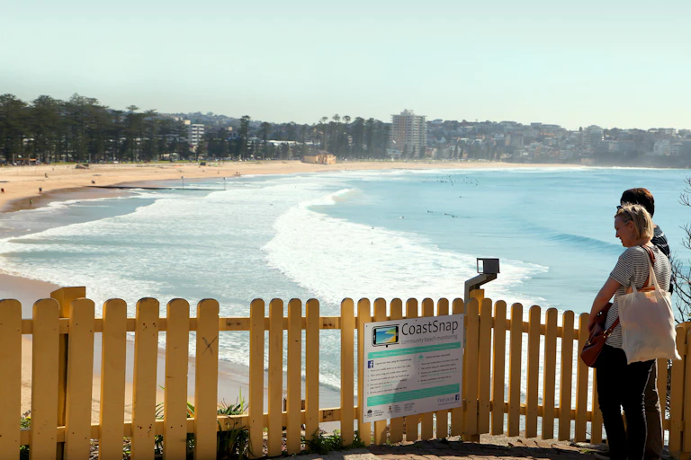 A couple approach a CoastSnap phone docking station with a view of a sandy beach