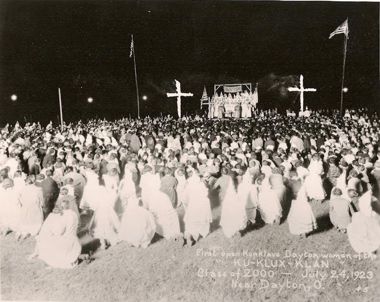 A black and white photograph shows a large crowd of people, many of them in white robes, crouched on the ground outside in front of a small stage with crosses on either side of it.