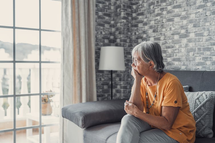 A senior woman sits on a couch looking out the window.