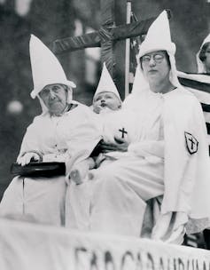 A black and white photograph of two women with a baby between them, all dressed in white robes and white hoods, with their faces showing.