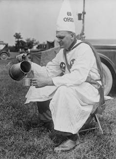 A man in a white robe and cap that says '69 Ohio' sits on a low stool outside pouring a drink from a pitcher.