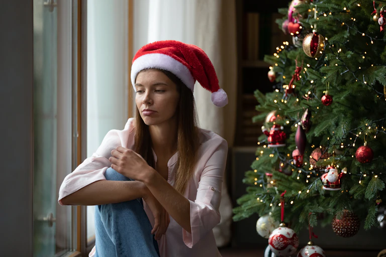 A young woman wearing a santa hat and sitting next to a Christmas tree looks sad.