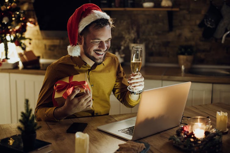 A man wearing a santa hat and holding a glass of champagne and a present smiles at someone he's speaking to on his laptop.