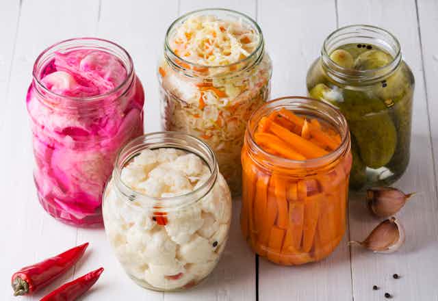 Clear jars full of pink pickled onions, cauliflower, carrots, cabbage, pickles in brine, as well as garlic and chilis. 