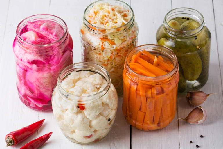 Clear jars full of pink pickled onions, cauliflower, carrots, cabbage, pickles in brine, as well as garlic and chilis.