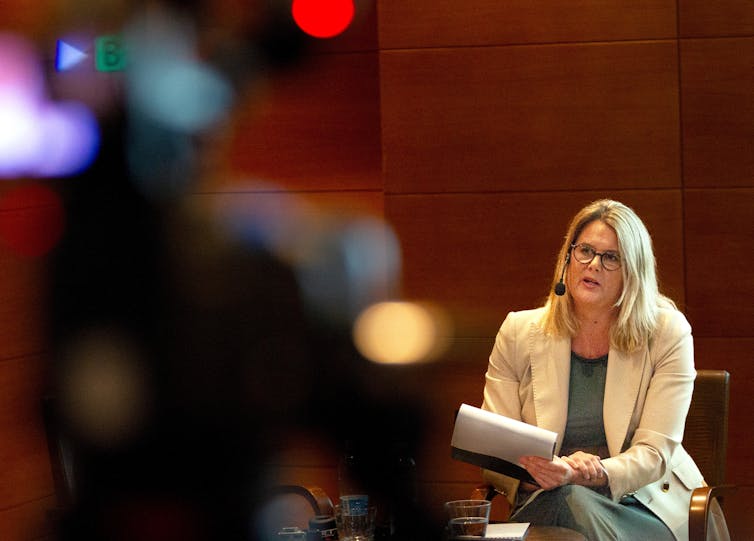 A person sits in a chair holding papers and speaking into a microphone.