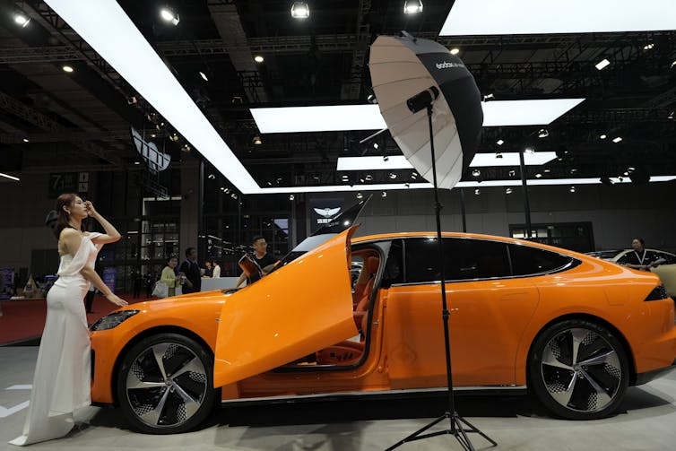 A woman in a white dress poses by a bright orange electric vehicle.