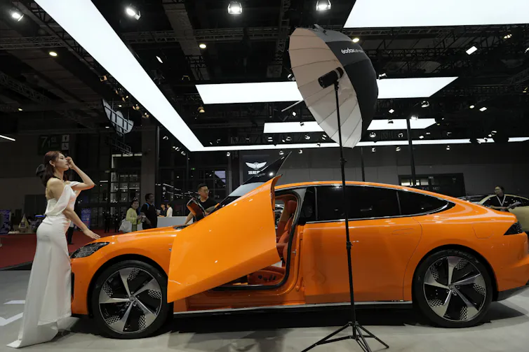 A woman in a white dress poses by a bright orange electric vehicle.