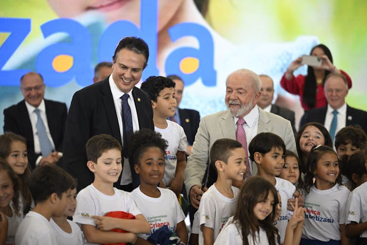 Several men in suits stand and smile behind a group of schoolchildren in white t-shirts.