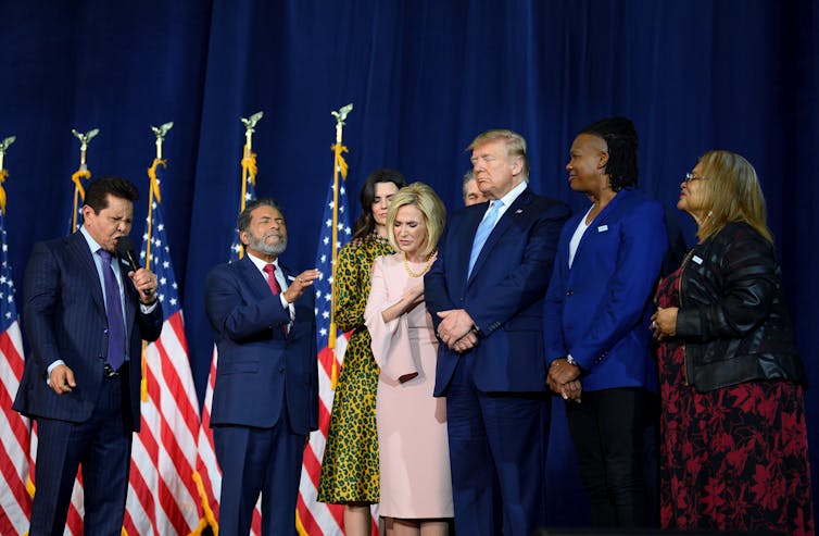 President Trump, in a navy blue suit, prays with his supporters standing on either side.
