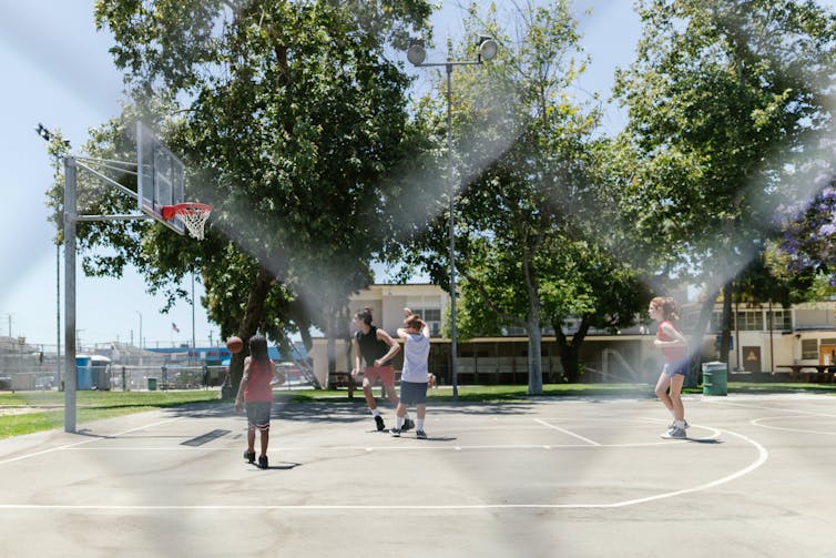 Young people play basketball against a backdrop of trees and low-rise buildings.