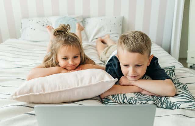 Two young children lie on a bed, watching a laptop. 
