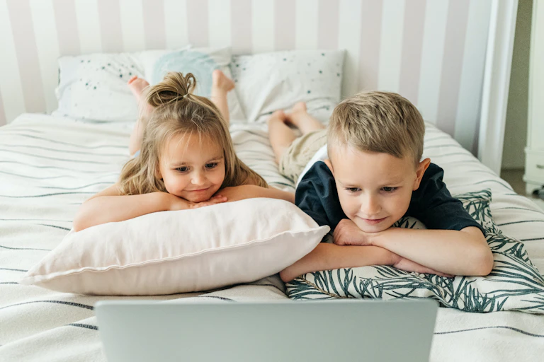 Two young children lie on a bed, watching a laptop.